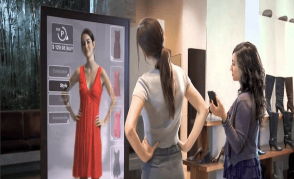 An image demonstrating the use of virtual mirrors. A person stands in front of a tall screen, wearing a t-shirt and jeans. The screen displays an image of them as if they are wearing a red dress. 